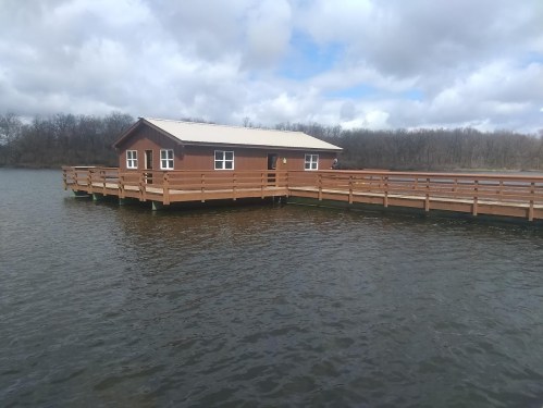 A wooden cabin on stilts over a calm body of water, surrounded by trees and cloudy skies.