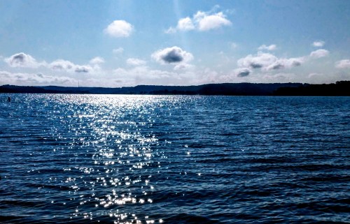 A serene lake scene with sparkling water under a bright blue sky and fluffy clouds in the distance.
