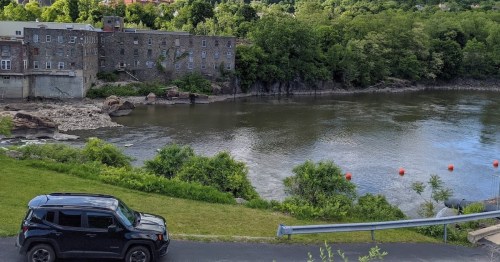 A black car parked near a riverbank with greenery and an old stone building in the background.