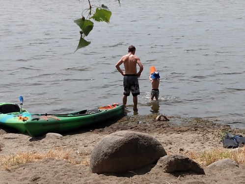 A man and a boy play by the water's edge, with a kayak nearby and trees framing the scene.