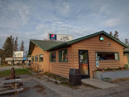 A rustic log cabin-style café with a green roof and Pepsi signage, surrounded by trees and outdoor seating.