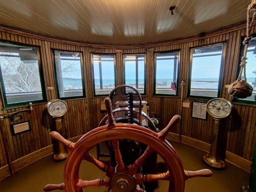 Interior of a lighthouse with a ship's wheel, gauges, and windows showing a view of the ocean and shoreline.