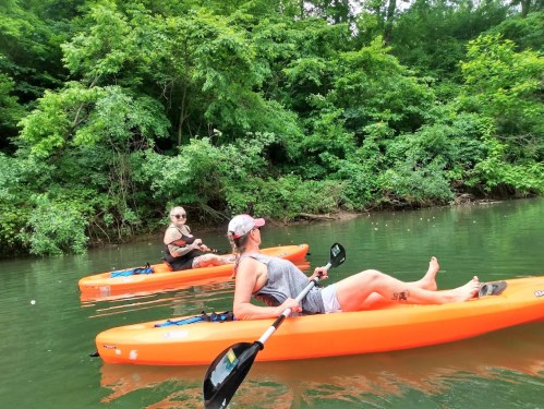 Two people in orange kayaks relax on a calm river surrounded by lush green trees.