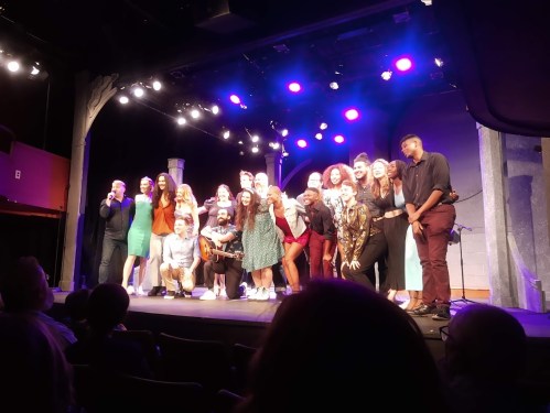 A diverse group of performers poses on stage, smiling under colorful lights during a theatrical event.
