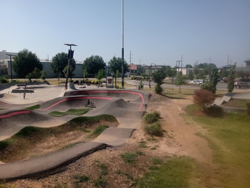 A sunny skate park with various ramps and paths, surrounded by greenery and a few people enjoying the space.