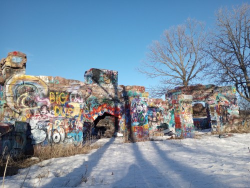 Colorful graffiti covers the ruins of a stone structure, surrounded by snow and bare trees under a clear blue sky.