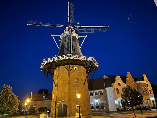 A windmill illuminated at night, with a clear blue sky and a crescent moon in the background. Nearby buildings are visible.