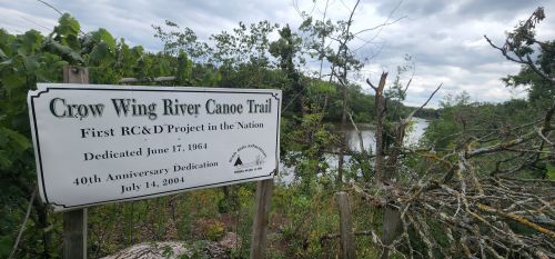 Sign for the Crow Wing River Canoe Trail, with a view of the river and trees in the background.