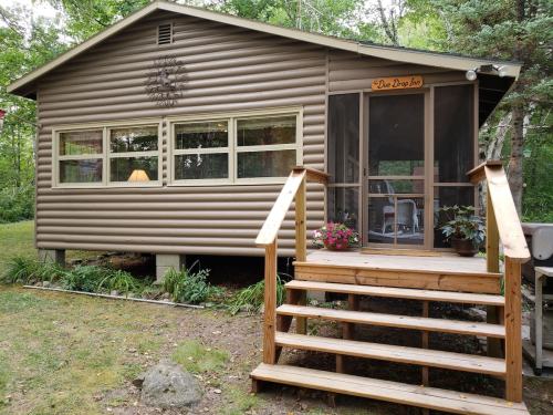 A cozy cabin with a wooden porch, surrounded by greenery and flowers, featuring large windows and a welcoming sign.