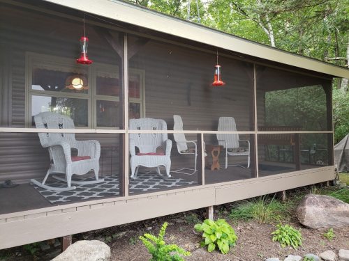 A screened porch with white wicker chairs, a rug, and hummingbird feeders, surrounded by greenery.