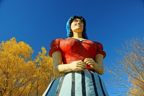A large, colorful statue of a woman with blue hair and a red top, set against a clear blue sky and autumn trees.