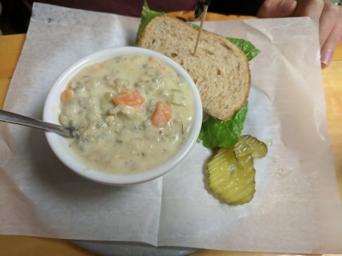 A bowl of creamy soup with vegetables, a slice of whole grain bread with lettuce, and a pickle on the side.
