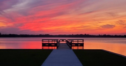 A serene lake at sunset, with vibrant pink and orange skies reflecting on the water and a wooden dock extending out.