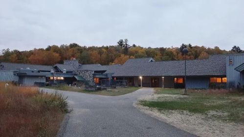 A modern building with a stone facade, surrounded by autumn trees, under a cloudy sky. Pathway leads to the entrance.