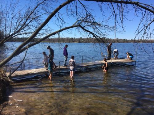A group of people wade in a lake near a wooden dock on a sunny day, with trees in the background.