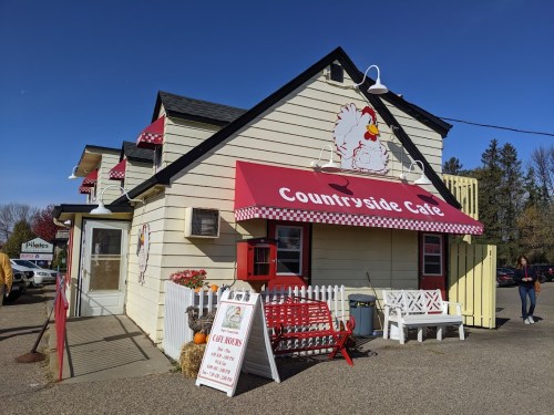 A charming café with a red awning featuring a chicken logo, surrounded by trees and outdoor seating.