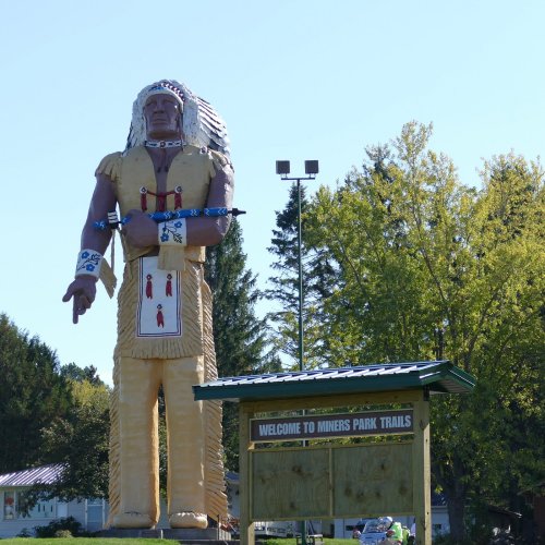 A large wooden statue of a Native American chief stands next to a sign that reads "Welcome to Miners Park Trails."