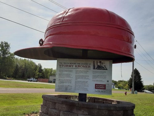 A large red hat sculpture with an informational sign about the legend of Stormy Kromer in a grassy area.