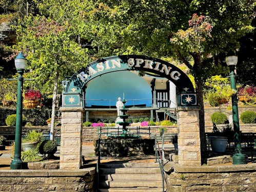 Archway sign reading "Basin Spring" with a fountain and landscaped gardens in a sunny park setting.