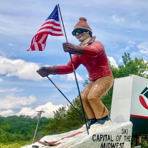 A large statue of a skier holding an American flag, wearing sunglasses and a red sweater, with a scenic background.