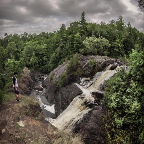 A person stands near a waterfall surrounded by lush green trees and rocky terrain under a cloudy sky.