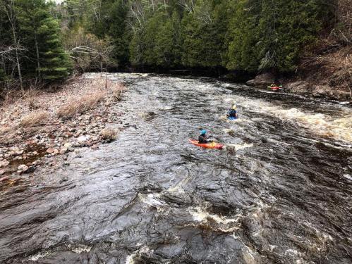 Two kayakers navigate a winding river surrounded by lush green trees and rocky banks.