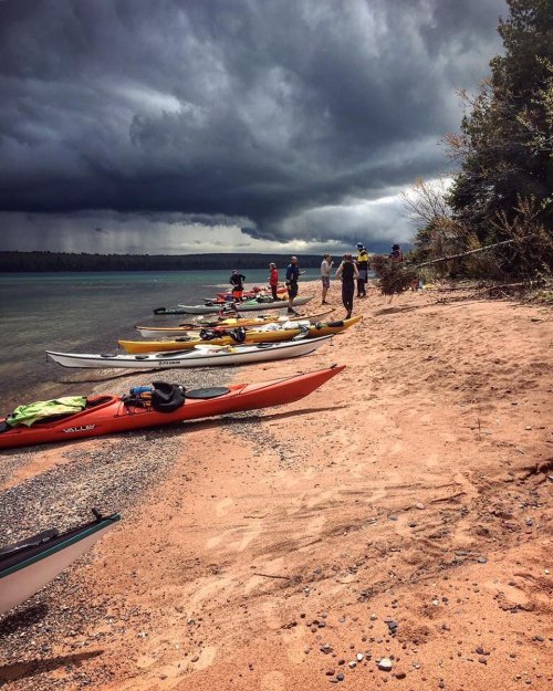 A sandy beach with kayaks lined up, people preparing for a trip under a cloudy sky.