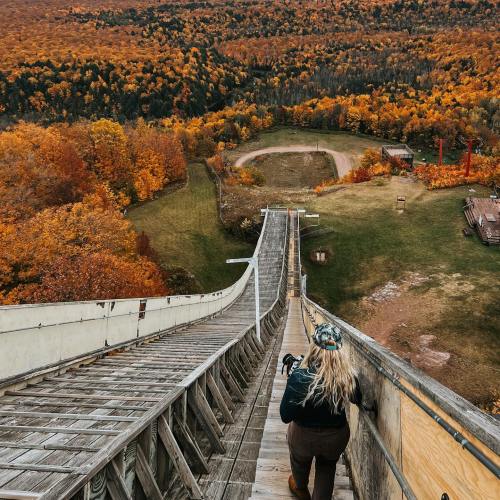 A person with long hair walks down a wooden ski jump, surrounded by vibrant autumn foliage and a scenic landscape.
