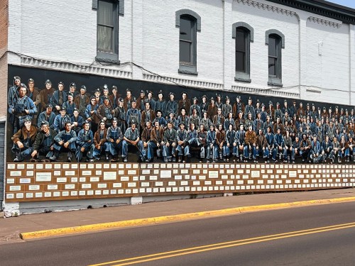 A large mural depicting numerous seated figures in blue and brown clothing, displayed on a building's exterior wall.