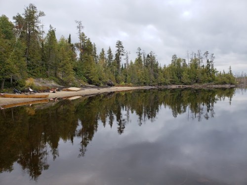A calm lake reflects trees and a cloudy sky, with a sandy shore and canoes visible along the water's edge.