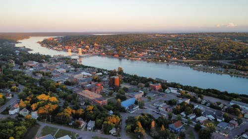 Aerial view of a riverside town with lush greenery, buildings, and a calm river reflecting the sunset.
