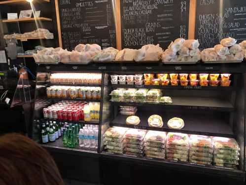 A display case filled with sandwiches, salads, drinks, and fresh fruit, alongside a chalkboard menu in a café.