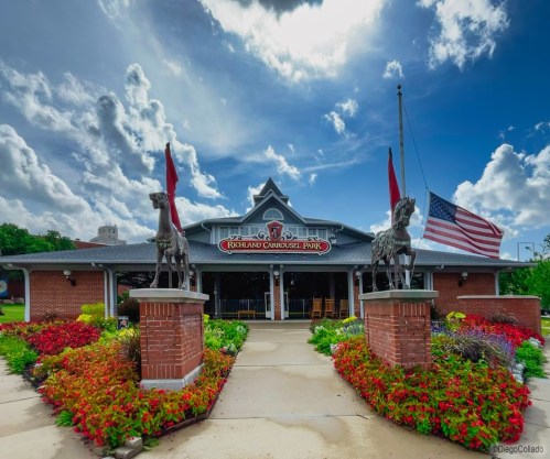 Entrance to Rockland Carousel Park, featuring horse statues, vibrant flowers, and a flag against a cloudy sky.