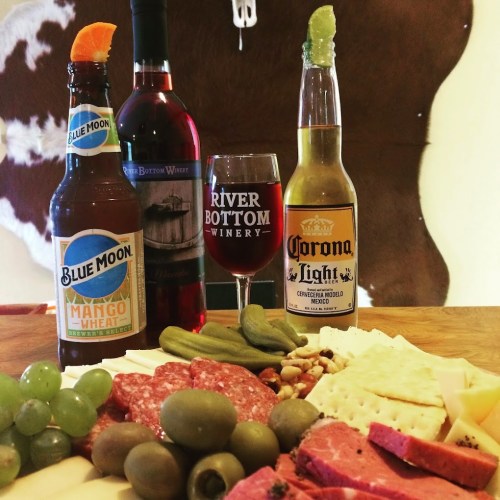 A wooden table with a cheese and charcuterie platter, featuring drinks from River Bottom Winery and two beer bottles.