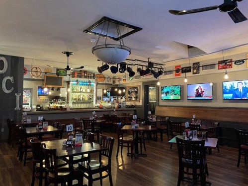 Interior of a restaurant with wooden tables, a bar, and multiple TVs displaying sports, decorated with memorabilia.