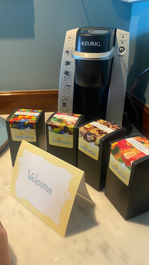 A Keurig coffee maker next to colorful snack boxes and a "Welcome" sign on a marble countertop.