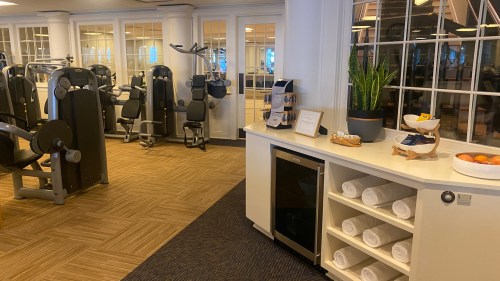 A modern gym with exercise equipment, a refreshment area, and neatly stacked towels in a bright, airy space.