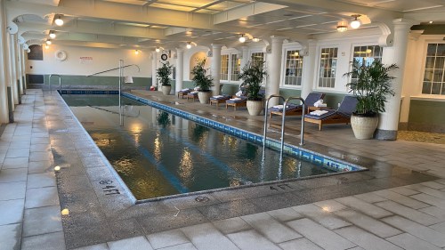 Indoor swimming pool area with lounge chairs, potted plants, and bright lighting. Water reflects the surrounding architecture.