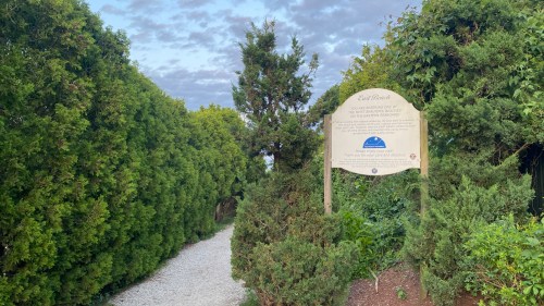 A gravel path leads through lush greenery to a sign for East Beach, with a cloudy sky above.