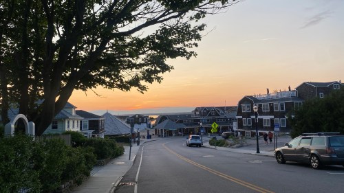 A scenic street lined with houses at sunset, with a view of the water in the distance and a car parked nearby.