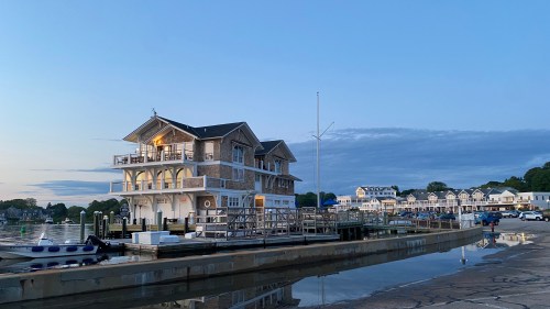 A waterfront building at dusk, with boats docked and a calm water reflection, surrounded by trees and distant houses.