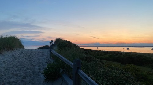 A sandy path leads to a serene beach at sunset, with boats on the water and lush greenery on either side.