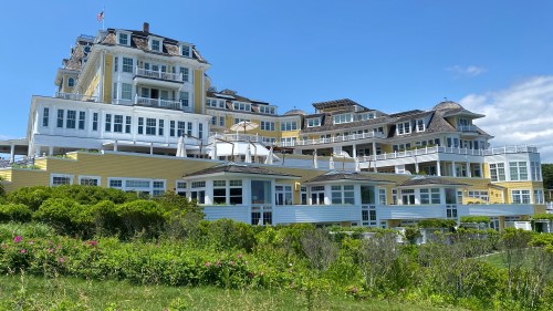 A large, elegant yellow hotel with multiple levels, surrounded by greenery and under a clear blue sky.