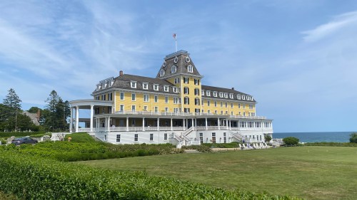 A large yellow historic hotel with a clock tower, surrounded by green lawns and overlooking the ocean under a blue sky.