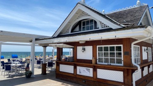 A beachside restaurant with a wooden exterior, outdoor seating, and ocean views under a clear blue sky.