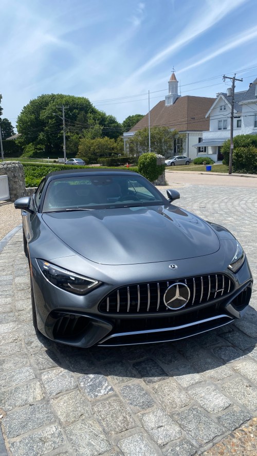 A sleek gray Mercedes-Benz sports car parked on a stone driveway with a residential area in the background.