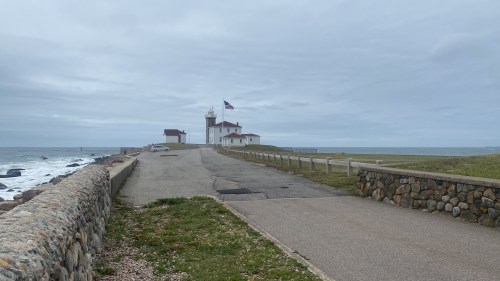 A lighthouse stands near the shore, with a road leading up to it and a cloudy sky overhead.