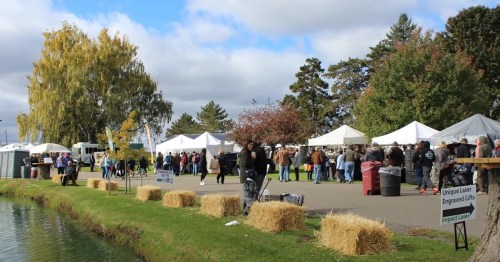 A bustling outdoor market with tents, people browsing, and a pond nearby, surrounded by trees and hay bales.