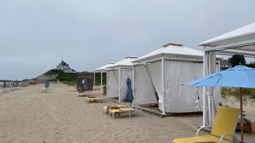 Beach cabanas with blue umbrellas and lounge chairs on sandy shore, with a cloudy sky and distant building in the background.