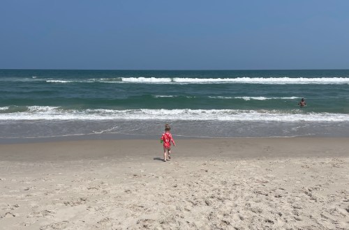 A child in a red swimsuit walks towards the ocean on a sandy beach, with gentle waves and a person swimming nearby.